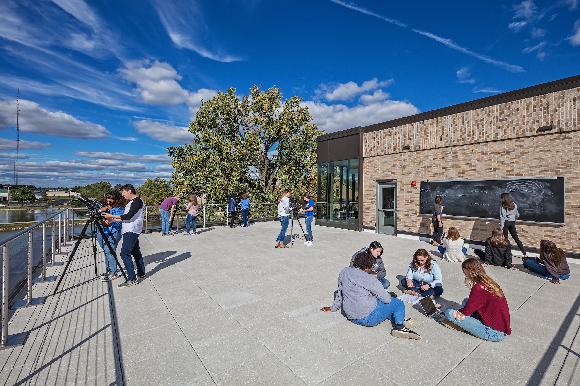 4 groups of students use telescopes while another group of 4 students sit and study and another group of students use the outdoor chalkboard