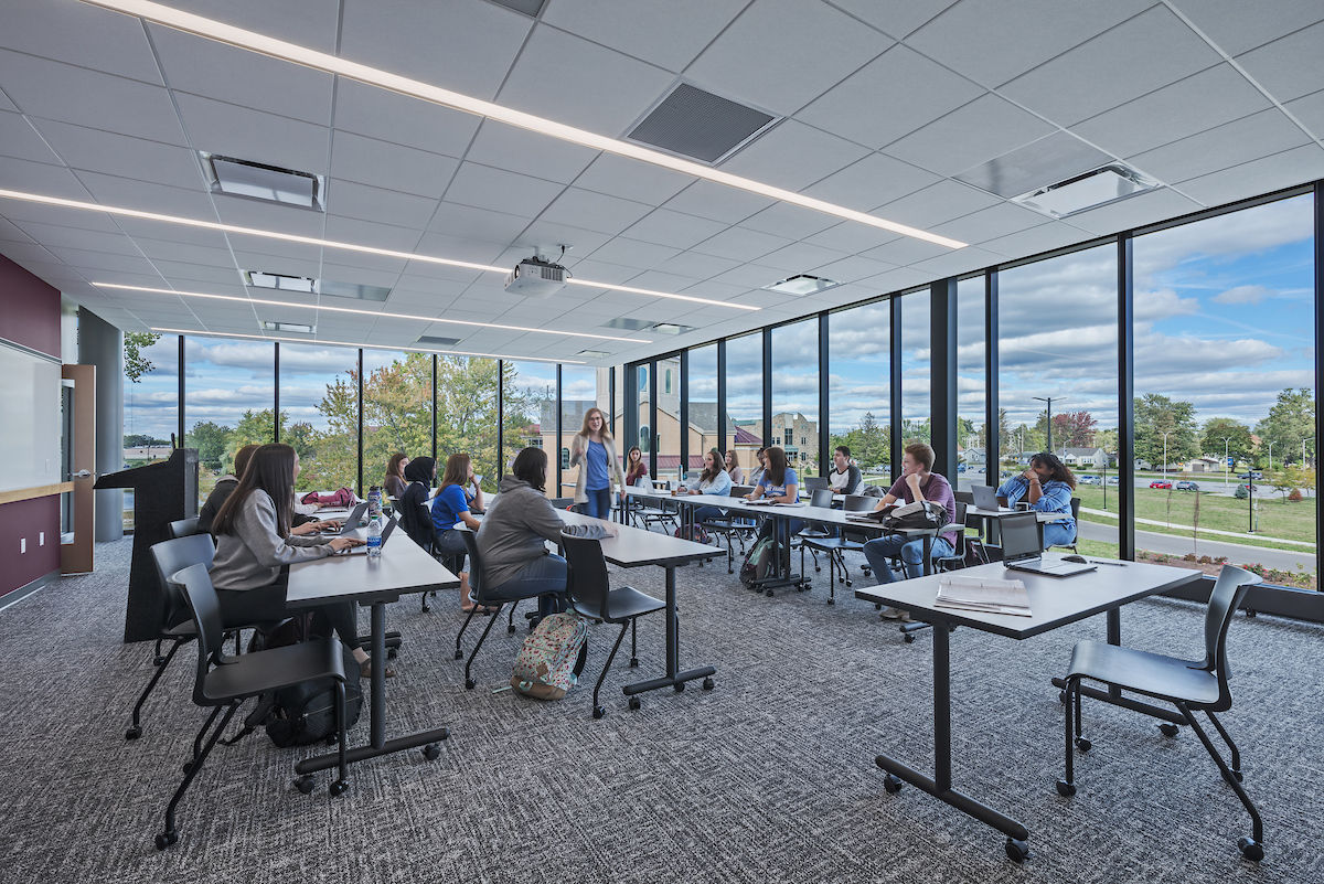 A professor teaches a large group of students in a classroom featuring floor to ceiling windows that overlook campus